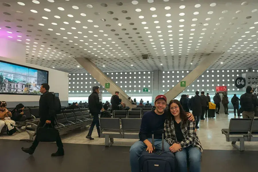 Pareja sonriente sentada en la zona de embarque de la Terminal 2 del Aeropuerto de la Ciudad de M&eacute;xico con su equipaje, esperando la salida de su vuelo